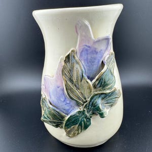 Puede incluir: Un jarrón de cerámica blanco con un diseño en relieve de flores moradas y verdes. El jarrón tiene un fondo redondeado y una abertura acampanada.