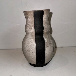 以下が含まれることがあります： 黒い縦縞が入った白い陶器の花瓶。花瓶は表面がざらざらしていて、丸みを帯びた形をしています。