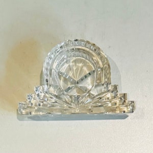 May include: A clear glass napkin ring with a decorative design. The ring is shaped like a circle with a raised centre.