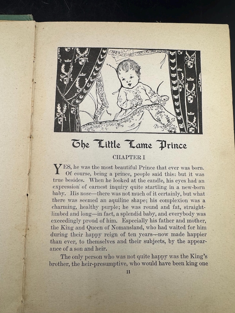 May include: A black and white illustration from "The Little Lame Prince" book. The image depicts a baby in a decorated bed, with text below. The baby is the central focus, rendered in a detailed drawing style.