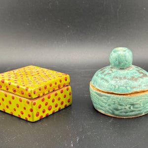 Peut inclure: Deux boîtes décoratives en céramique. L'une est carrée, jaune avec des pois rouges. L'autre est ronde, turquoise avec une surface texturée et un petit bouton sur le dessus. Les deux sont sur une surface sombre.