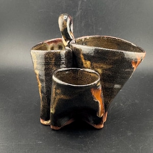 May include: A handcrafted ceramic utensil holder with a distinctive design. It has three compartments of different sizes, with a handle on the largest. The colour scheme includes black, brown, and orange, with a textured finish. Suitable for kitchen decor.