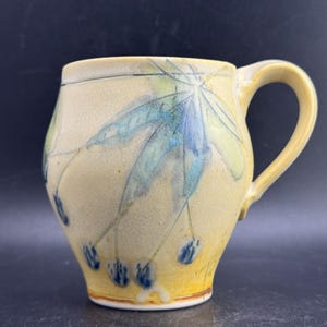 May include: A ceramic mug with a light yellow, speckled glaze featuring a hand-painted blue and green leaf design. The mug has a rounded body, a wide rim, and a curved handle on the right side. The base is slightly tapered with a white rim.