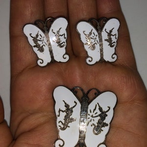 May include: A set of three silver butterfly earrings with white enamel and a detailed design of dancing figures.