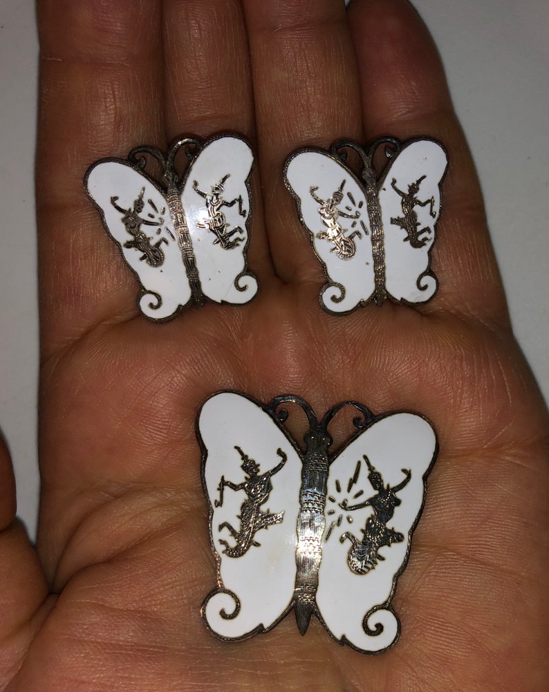 May include: A set of three silver butterfly earrings with white enamel and a detailed design of dancing figures.