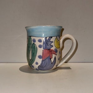 Puede incluir: Una taza de cerámica con un diseño azul, blanco y verde. La taza presenta un lobo de dibujos animados con un pañuelo rojo y un cactus.