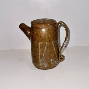 Può includere: Una piccola teiera in ceramica marrone con un coperchio e un manico. La teiera ha una glassa bianca con striature marroni.