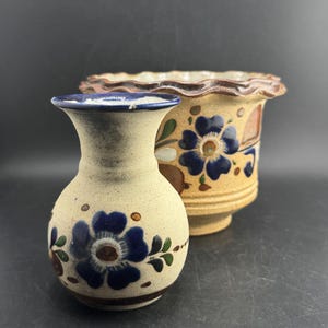 Puede incluir: Un jarrón pequeño y redondo de cerámica beige con borde azul y motivos florales azules pintados a mano, situado frente a una maceta de cerámica beige más grande y ancha con borde festoneado y diseños de flores azules y hojas marrones.