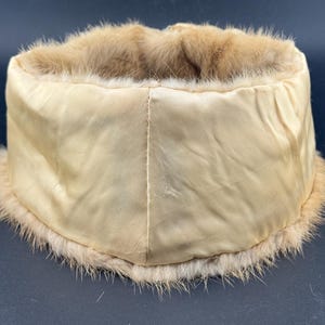 May include: A vintage hat featuring a cream satin exterior and a light brown fur trim. The hat has a cylindrical shape. The fur trim is soft and fluffy, adding a touch of luxury. The hat's design suggests it is from a bygone era, with a classic aesthetic.