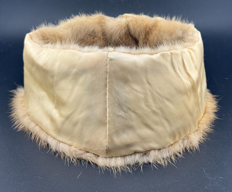 May include: A vintage hat featuring a cream satin exterior and a light brown fur trim. The hat has a cylindrical shape. The fur trim is soft and fluffy, adding a touch of luxury. The hat's design suggests it is from a bygone era, with a classic aesthetic.
