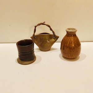 May include: Three small ceramic pots in different shapes and colors. A brown pot with a woven handle, a brown pot with a wide mouth, and a brown pot with a narrow mouth.