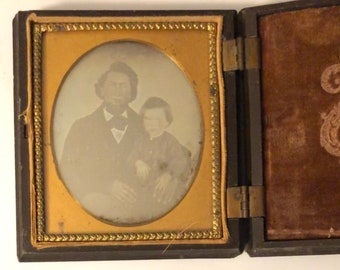 May include: A vintage tintype photograph of a man and a child in a wooden frame. The man is wearing a dark suit and a hat. The child is wearing a dark dress. The photograph is in a black and gold frame.
