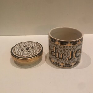 May include: A white ceramic jar with a black and gold mosaic design. The jar has the word "du jour" written in black letters on the front. The jar has a matching lid.