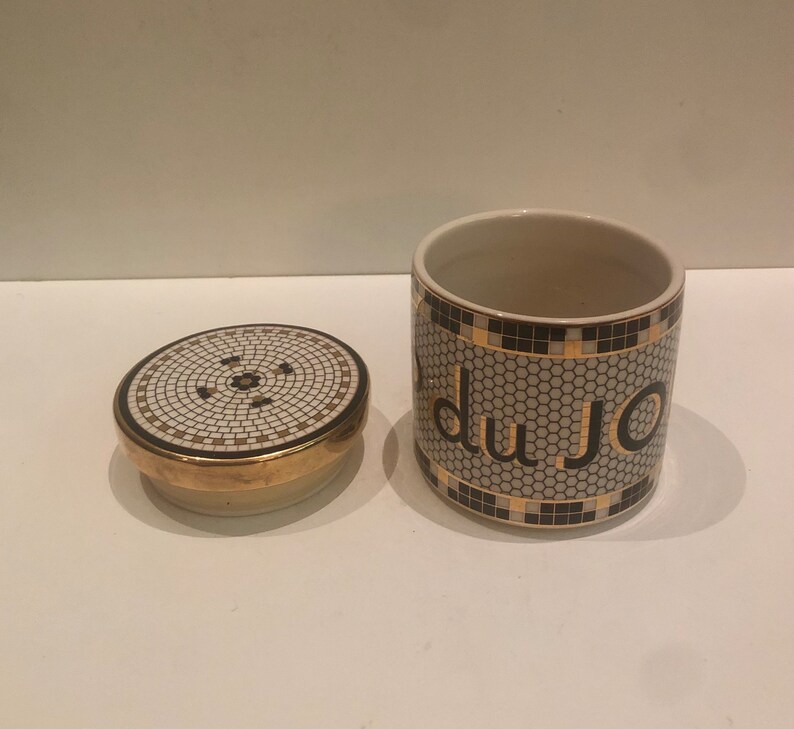 May include: A white ceramic jar with a black and gold mosaic design. The jar has the word "du jour" written in black letters on the front. The jar has a matching lid.