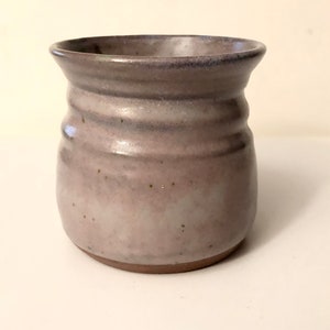 Op de afbeelding: Een kleine, handgegooide keramische pot met een lichtpaarse glazuur. De pot heeft een rond lichaam en een licht uitlopende rand.
