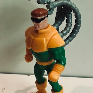 May include: A green and yellow action figure of Doctor Octopus, a supervillain from the Spider-Man comics. The figure has four mechanical arms attached to its back.