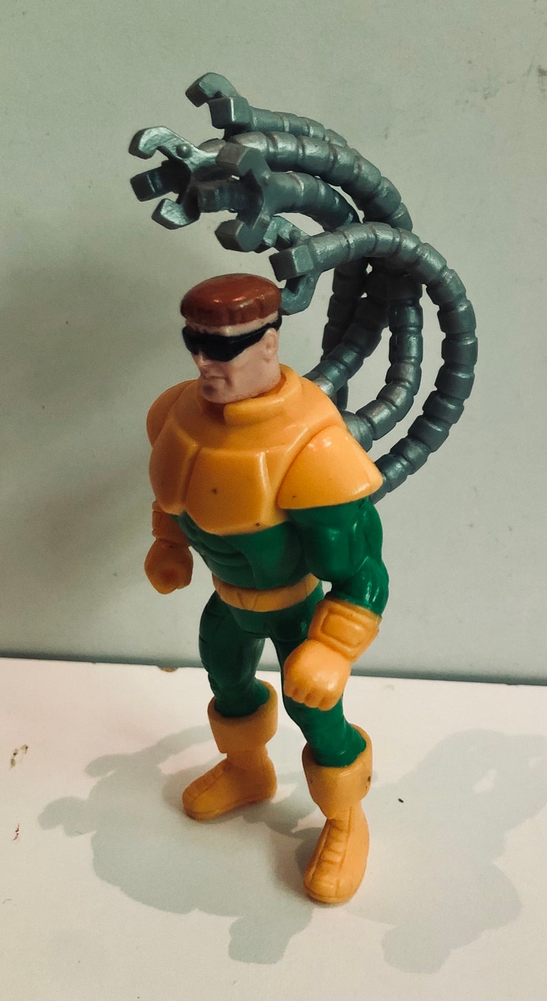 May include: A green and yellow action figure of Doctor Octopus, a supervillain from the Spider-Man comics. The figure has four mechanical arms attached to its back.