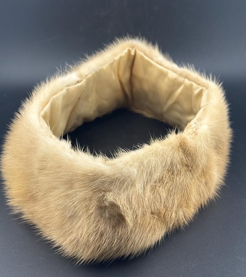 May include: A light brown fur collar with a soft, beige interior lining. The collar is circular and appears to be made of a soft, plush material. The fur has a natural, textured appearance, and the collar is set against a dark background.
