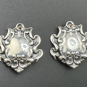 May include: Two ornate, silver-toned pendants with a shield-like center and intricate scrollwork. Each pendant has a small loop at the top for hanging. The pendants are displayed on a dark gray surface.