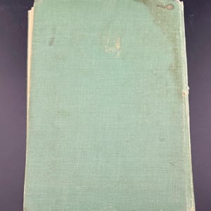 May include: A vintage book with a faded, pale green cover. The cover has a textured appearance and shows signs of wear, including discoloration and small spots. The book's edges are slightly worn, suggesting age and use. The overall aesthetic is one of a well-loved, antique item.