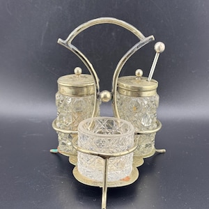 May include: A silver-tone condiment set with three glass containers. The containers have a cut-glass design and are held in place by a metal stand with a handle.