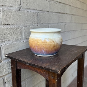 May include: A small, white ceramic planter with a brown, orange, and purple glaze. The planter is sitting on a dark brown wooden table.