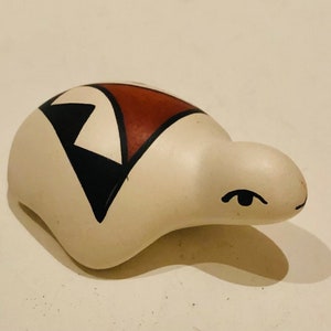 Puede incluir: Una figurita de tortuga de cerámica blanca con diseños geométricos negros y marrones en su caparazón. La tortuga tiene un ojo negro simple y una pequeña boca.