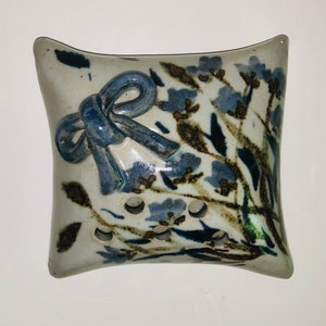 以下が含まれることがあります： 青い花柄と青いリボンが付いた白い陶器の枕。枕の下部に排水用の穴が開いています。