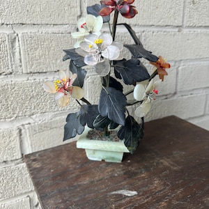May include: A decorative floral arrangement featuring a variety of colorful, translucent flowers with yellow centers and dark green leaves. The flowers are set in a light green square pot, resting on a dark wooden table. The background is a white brick wall.
