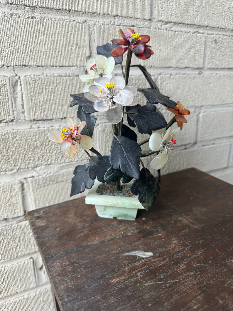 May include: A decorative floral arrangement featuring a variety of colorful, translucent flowers with yellow centers and dark green leaves. The flowers are set in a light green square pot, resting on a dark wooden table. The background is a white brick wall.