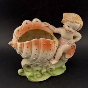 Puede incluir: Una maceta de cerámica vintage con forma de concha, con una figura de querubín. La concha es blanca con detalles en naranja y puntos dorados. El querubín tiene cabello rubio y una faja rosa. La base es verde.