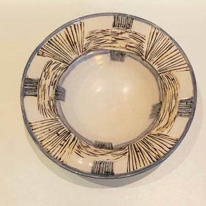 Puede incluir: Un cuenco de cerámica blanco con un borde azul y un diseño decorativo. El borde presenta un diseño repetitivo de líneas negras y curvas. El interior del cuenco es de color blanco roto liso, creando un contraste con el borde estampado.