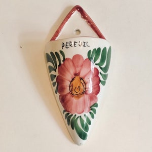 May include: White ceramic wall planter with a pink flower and green leaves. The planter has a triangular shape and says "Perejil" on the front.