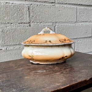 May include: A vintage ceramic covered dish with a cream base and a brown glaze. The dish has a gold trim and floral design.
