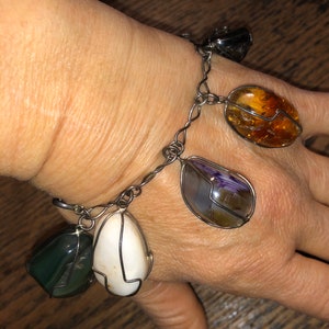 Pode incluir: Um bracelete de corrente de prata com cinco pingentes de pedras preciosas diferentes. Os pingentes s&atilde;o uma pedra cinza escura, uma pedra branca, uma pedra roxa e marrom, uma pedra transparente e uma pedra &acirc;mbar.