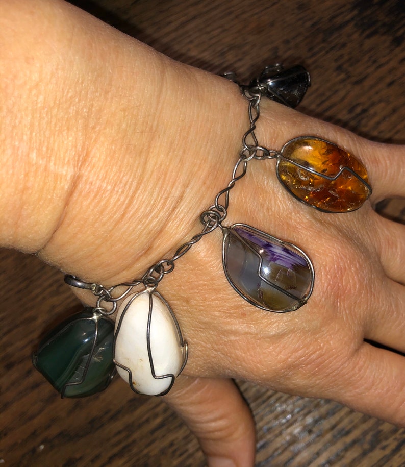 Pode incluir: Um bracelete de corrente de prata com cinco pingentes de pedras preciosas diferentes. Os pingentes s&atilde;o uma pedra cinza escura, uma pedra branca, uma pedra roxa e marrom, uma pedra transparente e uma pedra &acirc;mbar.