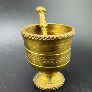 Könnte beinhalten: Ein goldfarbener Mörser und Stößel mit einem dekorativen Design. Der Mörser hat einen runden Boden und eine breite, flache Schale. Der Stößel ist ein langer, dünner Stab mit einem abgerundeten Ende.