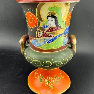 Puede incluir: Jarrón decorativo de cerámica pintado a mano con la parte superior acampanada y base de pedestal. El jarrón presenta una ilustración detallada de una figura con halo, flores y detalles dorados. La paleta de colores incluye negro, naranja, verde y dorado.