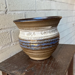 May include: A brown and white ceramic pot with blue and gold stripes. The pot has the text "1991 WXX Chickama" inscribed on the side.