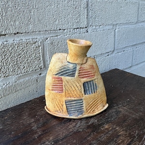 May include: A small, hand-crafted ceramic vase with a geometric pattern in shades of yellow, red, blue, and brown. The vase has a wide base and a narrow neck.
