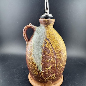 May include: A handcrafted ceramic oil bottle with a stainless steel spout. The bottle features a handle and a decorative design in shades of brown, yellow, and green. It sits on a matching ceramic base, perfect for kitchen use.