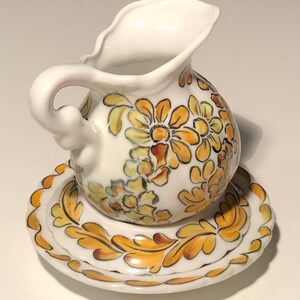 May include: A small white porcelain creamer with a floral design in shades of yellow and brown. The creamer has a handle and sits on a matching saucer.