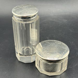 May include: Two antique glass jars with silver-colored lids. The taller jar is cylindrical with vertical facets. The smaller jar is a round, faceted shape. Both lids have an ornate floral design.