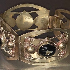 Puede incluir: Un brazalete vintage dorado con bisagras y detalles ornamentados. El brazalete presenta piedras negras de forma ovalada y recortes decorativos. El dise&ntilde;o incluye peque&ntilde;os acentos redondos y filigranas met&aacute;licas, creando una est&eacute;tica antigua.