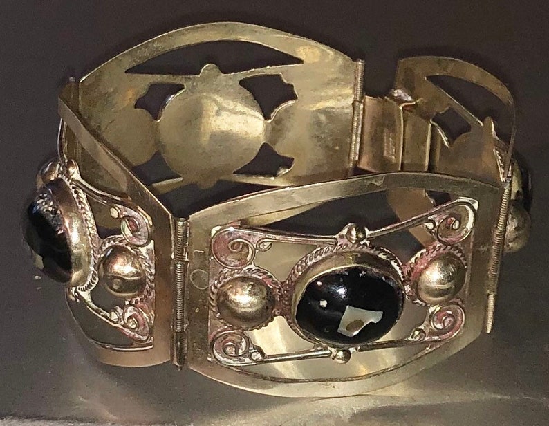 Puede incluir: Un brazalete vintage dorado con bisagras y detalles ornamentados. El brazalete presenta piedras negras de forma ovalada y recortes decorativos. El dise&ntilde;o incluye peque&ntilde;os acentos redondos y filigranas met&aacute;licas, creando una est&eacute;tica antigua.
