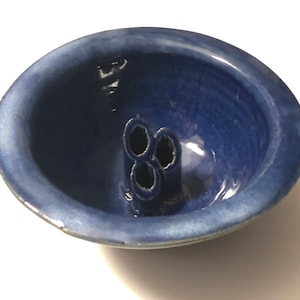 Può includere: Una piccola ciotola in ceramica blu con tre fori sul fondo. La ciotola è smaltata e ha una leggera lucentezza.