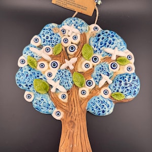 May include: A ceramic tree wall hanging decorated with blue and white evil eye charms and white doves. The tree is brown and has a textured surface. The text on the tag reads "AĞAÇ GÜÇTÜR BEREKETTİR İNSANLARIN İBRİYLE VE DOĞANIN İNSANLARLA BAĞINI SEMBOLİZE EDER. KORUYUCU VE ARINDIRICI NİTELİĞİNE SAHİPTİR. ALEDİR,  SOYUTLARIN VE KORKULARIN ÖNLEYİCİSİDİR."