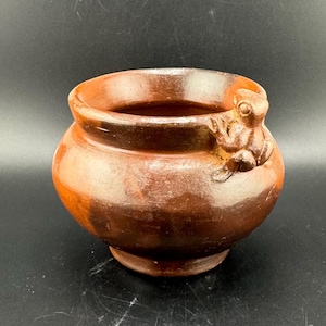 Puede incluir: Una maceta de terracota con un asa de rana. La maceta es marrón y tiene una forma redondeada. La rana es marrón y tiene una superficie texturizada.