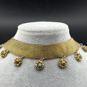 May include: A gold-toned choker necklace with a woven band and dangling floral pendants. The pendants feature small, colorful gemstones, including blue, red, and turquoise. The necklace is displayed on a gray mannequin head against a dark background.