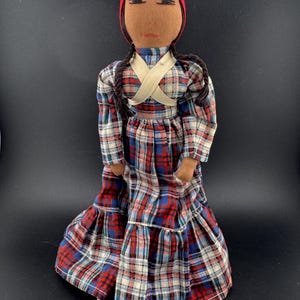Może przedstawiać: Ręcznie robiona lalka z materiałową twarzą, w czerwonym nakryciu głowy i sukience w kratę w kolorze czerwonym, niebieskim i białym. Lalka ma warkocze i kremowy pasek na ramię.